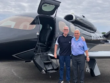Two men stand arm in arm on the tarmac beside a black light jet with its airstair door open