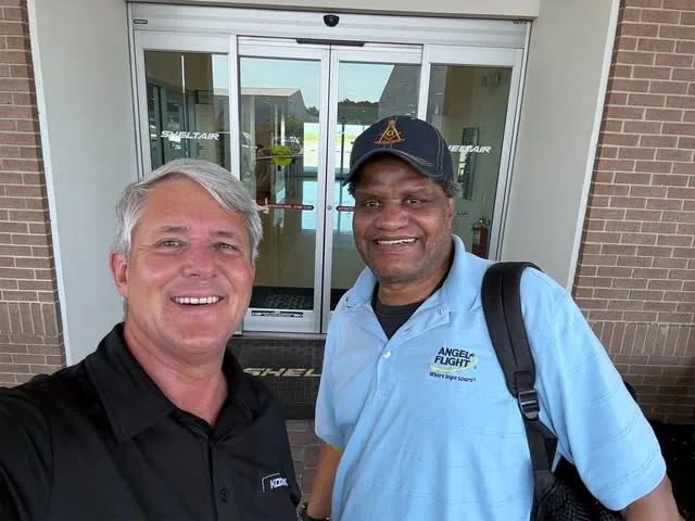 Two smiling men stand outside a building entrance, one wearing an Angel Flight polo shirt and carrying a shoulder bag