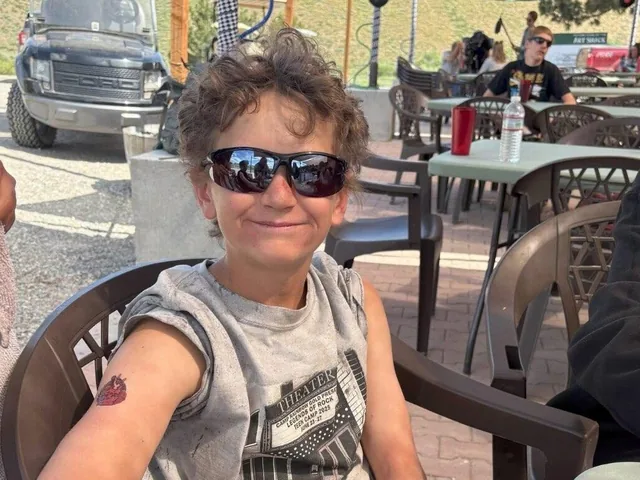 A child in sunglasses sits at an outdoor table at summer camp, smiling toward the camera