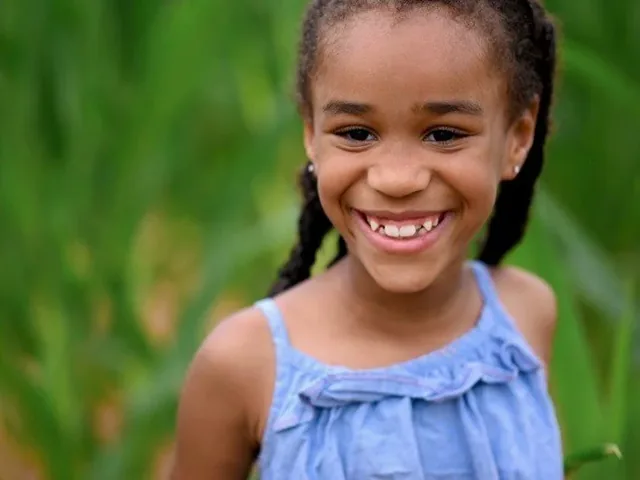 Alana, a young girl in a blue sundress, smiles brightly while standing in tall green grass