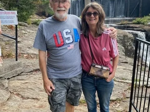An older couple stand together on a rocky overlook beside a safety railing