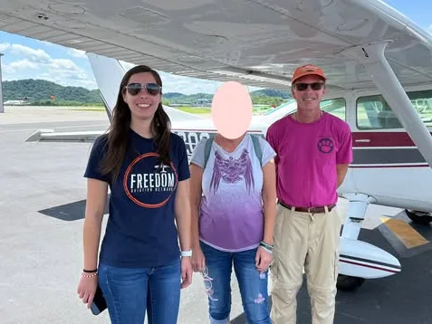 Three people stand in front of a high-wing Cessna on the ramp; one person's face is blurred for privacy