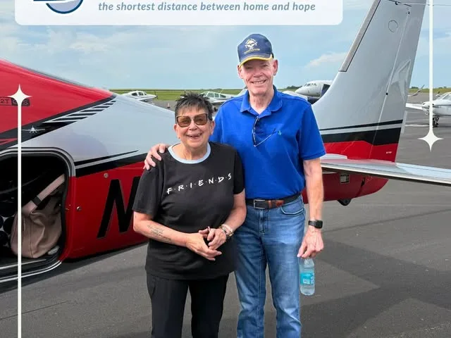 A passenger and a volunteer pilot stand together beside a red and white single-engine aircraft