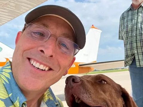 A volunteer pilot takes a smiling selfie on the ramp beside Brownlee, a brown Chesapeake Bay Retriever, under the wing of a small aircraft