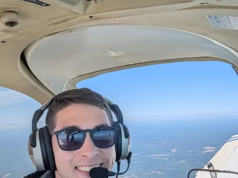 A volunteer pilot wearing sunglasses and a headset smiles in the cockpit with land visible far below