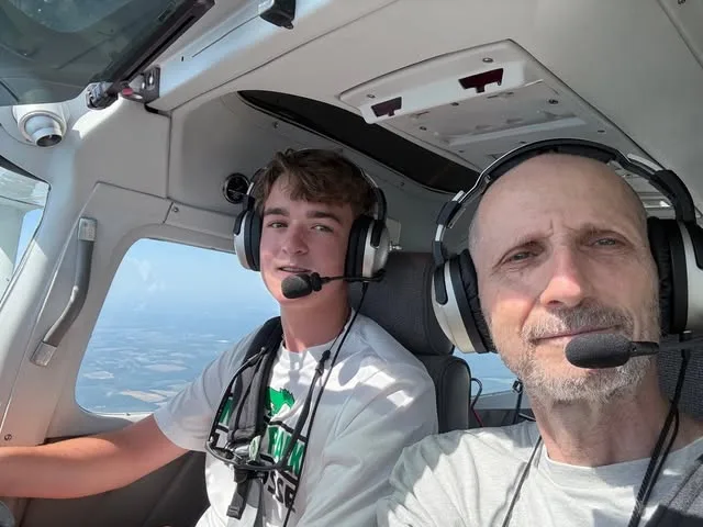 A teenage student and a volunteer pilot mentor wearing headsets sit side by side in a cockpit in flight