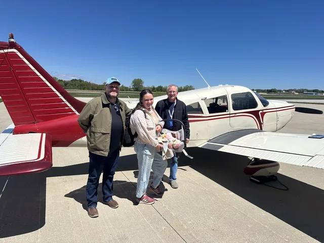 Two volunteers and a parent holding an infant stand on the ramp beside a red and white Piper